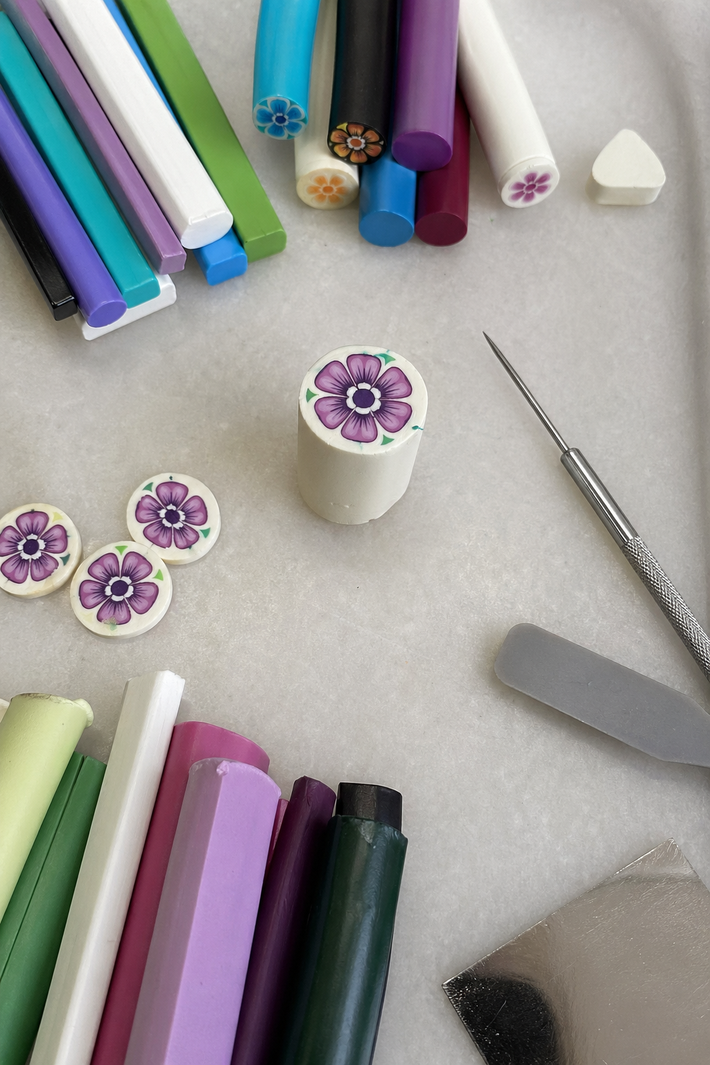 A photorealistic top-down view of a workspace with polymer clay canes in various colors, a needle tool, a silicone spatula, and a half-finished floral cane. Alt: Advanced polymer clay sculpting techniques workspace with canes and tools.