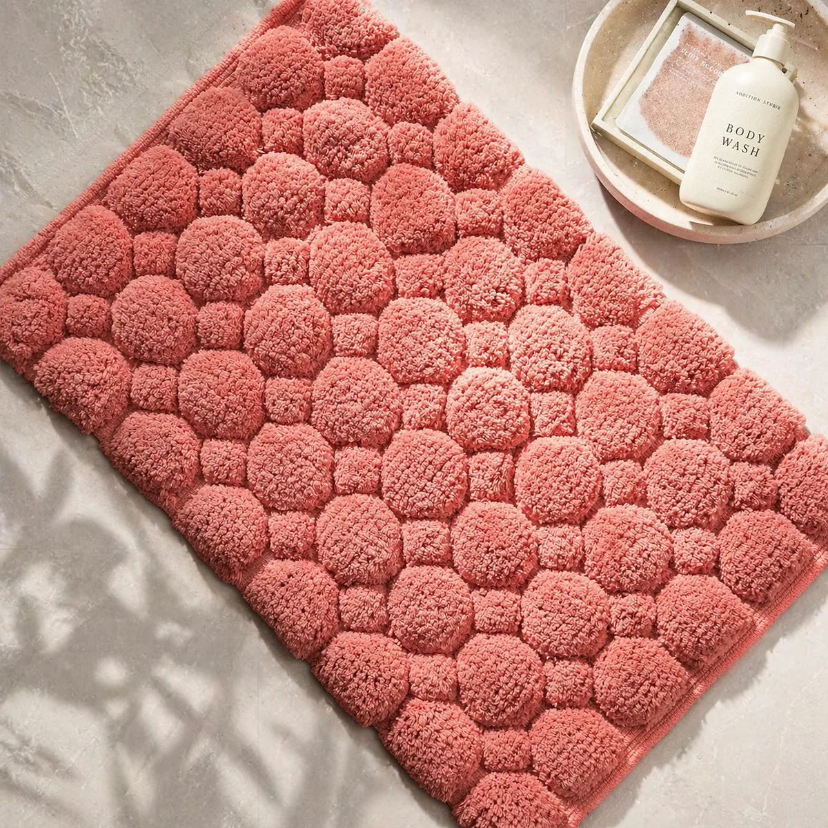 Coral woven cotton bath mat with a raised pebble stone texture next to a tray of bath products