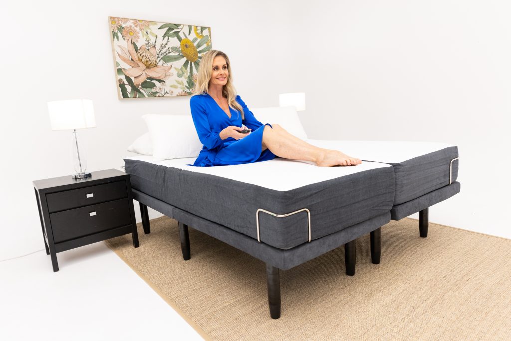 A realistic bedroom scene showing a modern adjustable bed base with a sleek remote control, a person reclining with feet
