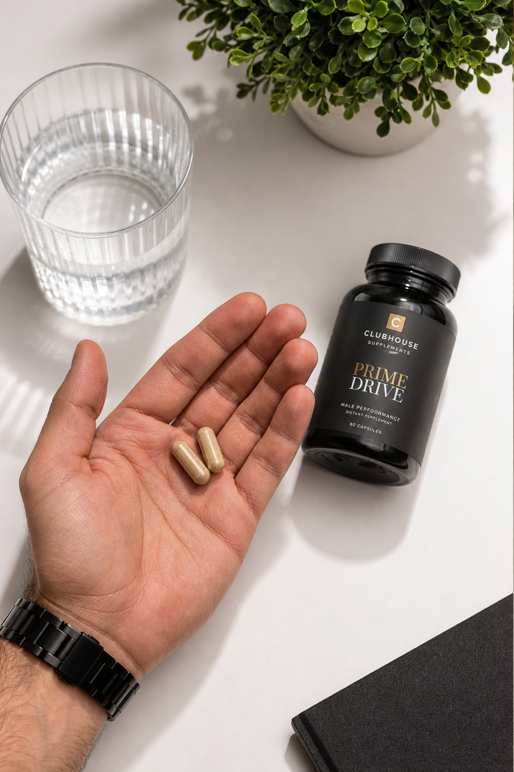 A photorealistic top-down view of a man's hand holding two supplement capsules over a clean white table, with a glass of water and a small plant in the background, conveying a health-focused lifestyle. Alt: man holding arousal supplement capsules healthy lifestyle