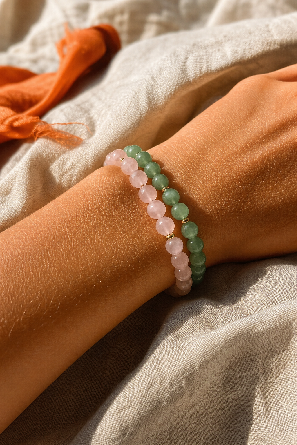 A photorealistic image of pink and green gemstone bracelets layered on a woman's wrist resting on a soft linen blanket in natural light. Alt: rose quartz and green aventurine love crystal bracelet combination.