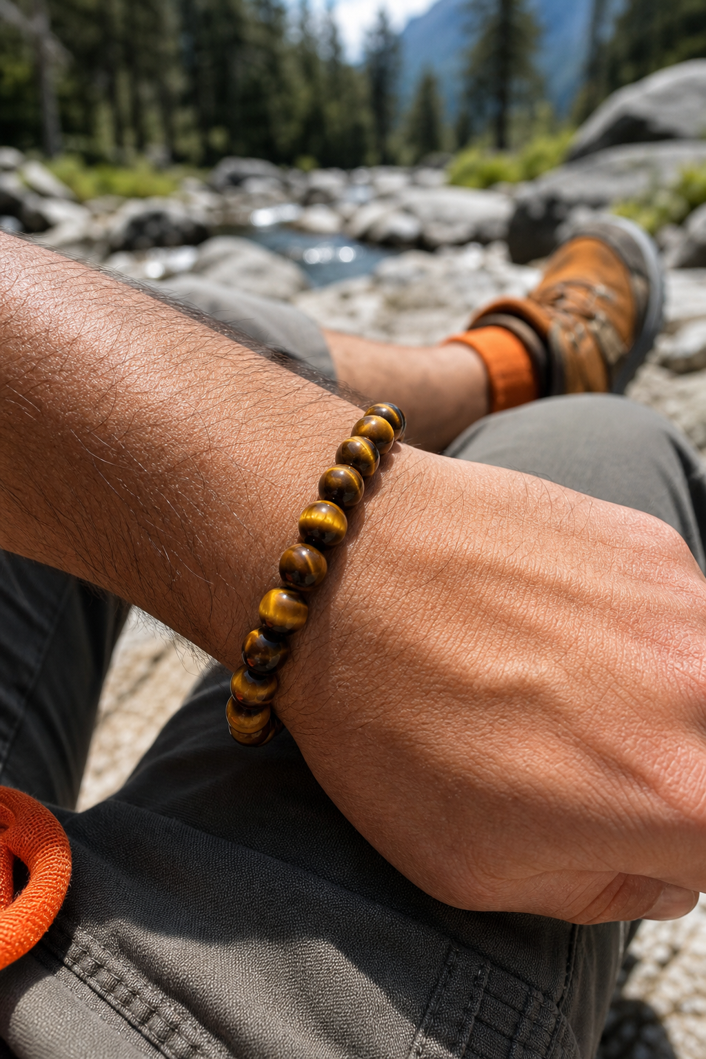 A close-up photorealistic image of a man's wrist wearing a golden-brown tiger eye beaded bracelet, with sunlight catching the chatoyant stripes. The background shows a natural outdoor setting with rocks and greenery. Alt: Man wearing a tiger eye crystal bracelet for confidence in a natural setting.