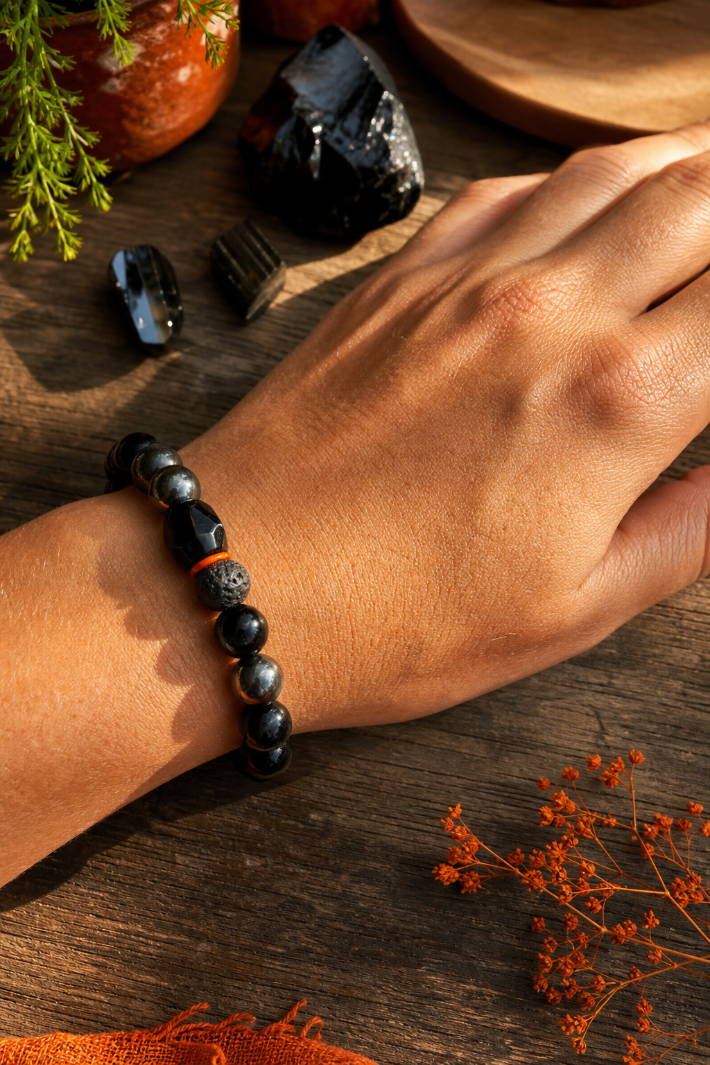 A photorealistic close-up of a hand adorned with a black tourmaline, obsidian, and hematite bracelet resting on a sunlit wooden table, with soft shadows and a calm, earthy background. Alt: Auras of Osiris triple protection crystal bracelet on a hand