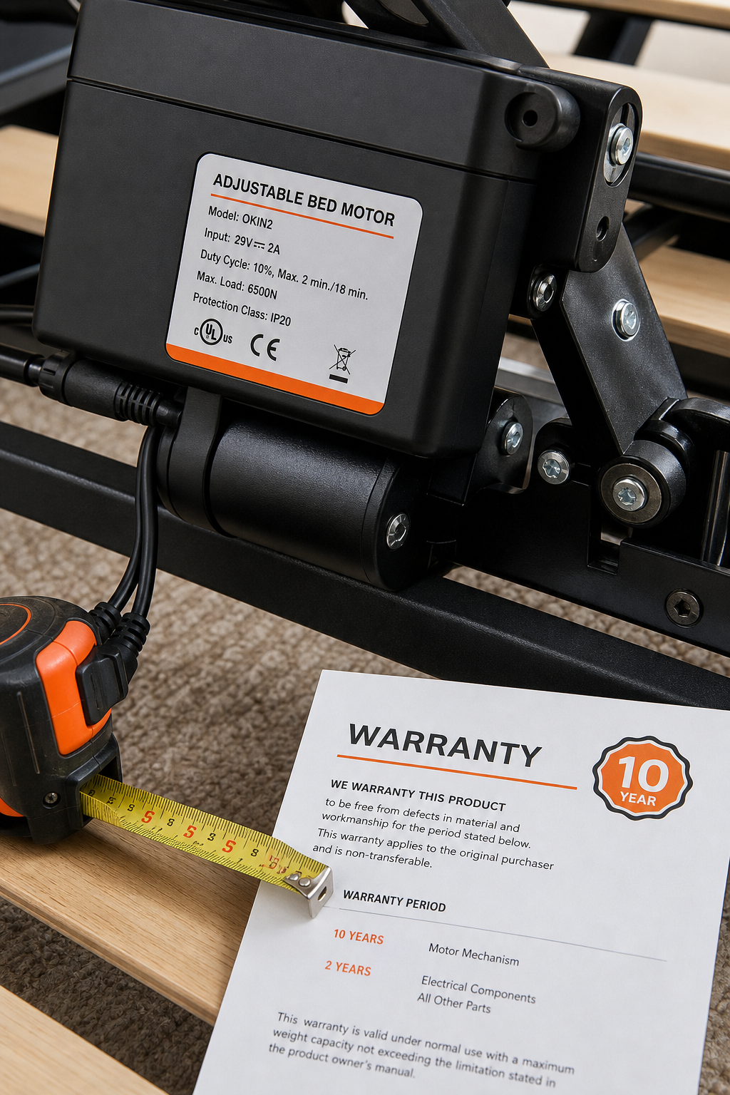 A close-up of an adjustable bed's motor and control unit, showing the label with specifications, with a tape measure and warranty paper nearby. Alt: Adjustable bed motor mechanism close-up with warranty details.