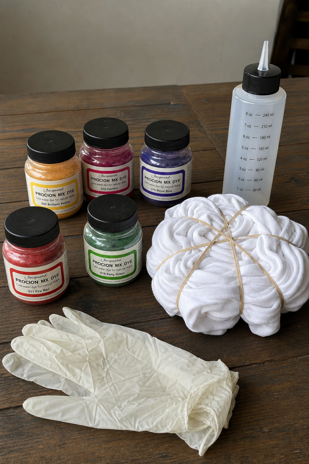A photorealistic scene of a workspace with several small jars of colorful dye powders, a white cotton t-shirt folded with rubber bands, and a pair of gloves, all arranged on a wooden table. Alt: Dyeing supplies including fiber-reactive dyes and a tied t-shirt for tie-dye.