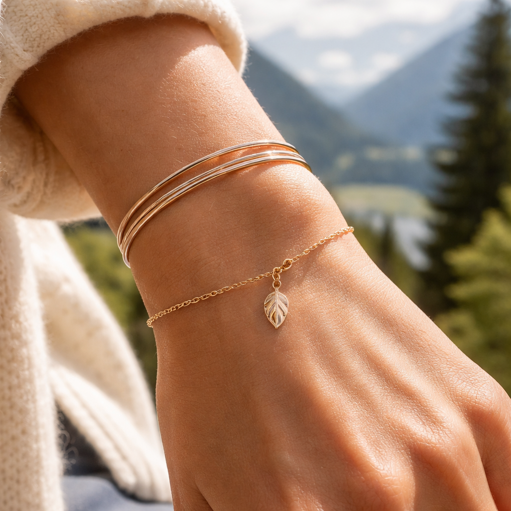 A photorealistic close‑up of a woman's wrist wearing thin mixed‑metal rings and a tiny leaf charm on a delicate gold chain, soft natural light, Swiss backdrop, realistic style.