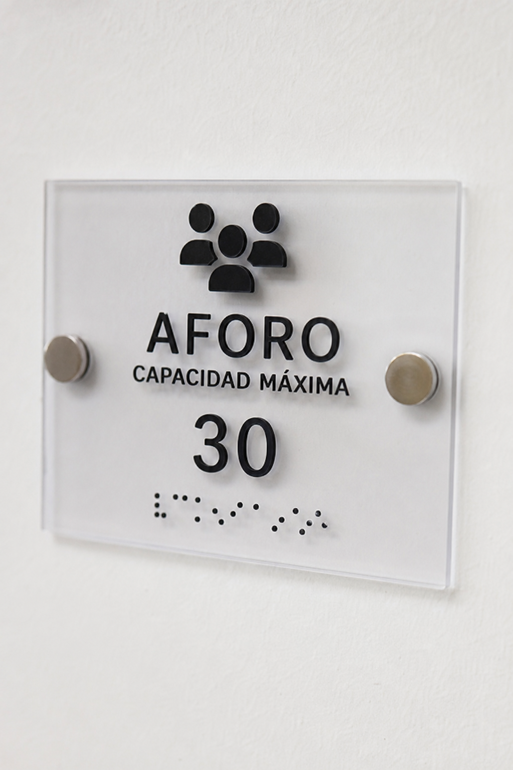 Realismo: Fotografía de un cartel de señalética braille en acrílico montado en una pared blanca, con texto en alto relieve y puntos braille visibles, iluminación suave, fondo limpio. Alt: Cartel de señalética braille en acrílico mostrando texto y braille en alto relieve.