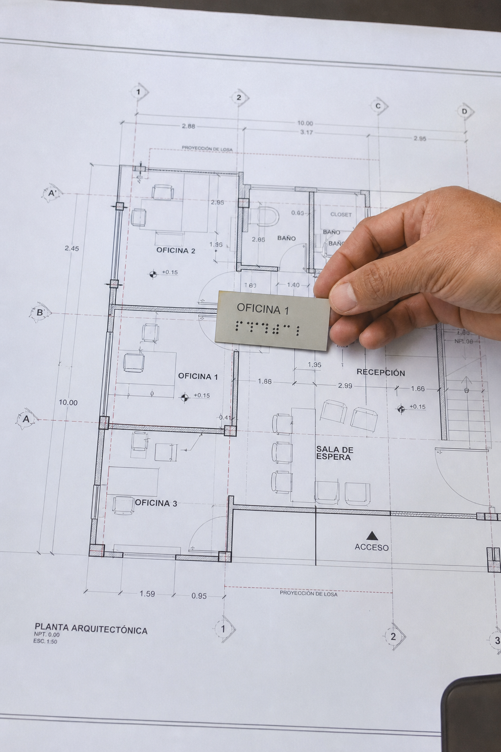 Realismo: Fotografía de un plano arquitectónico sobre una mesa de dibujo, con una mano colocando una placa braille de muestra. Alt: Plano arquitectónico con muestra de señalética braille para verificar ubicación.