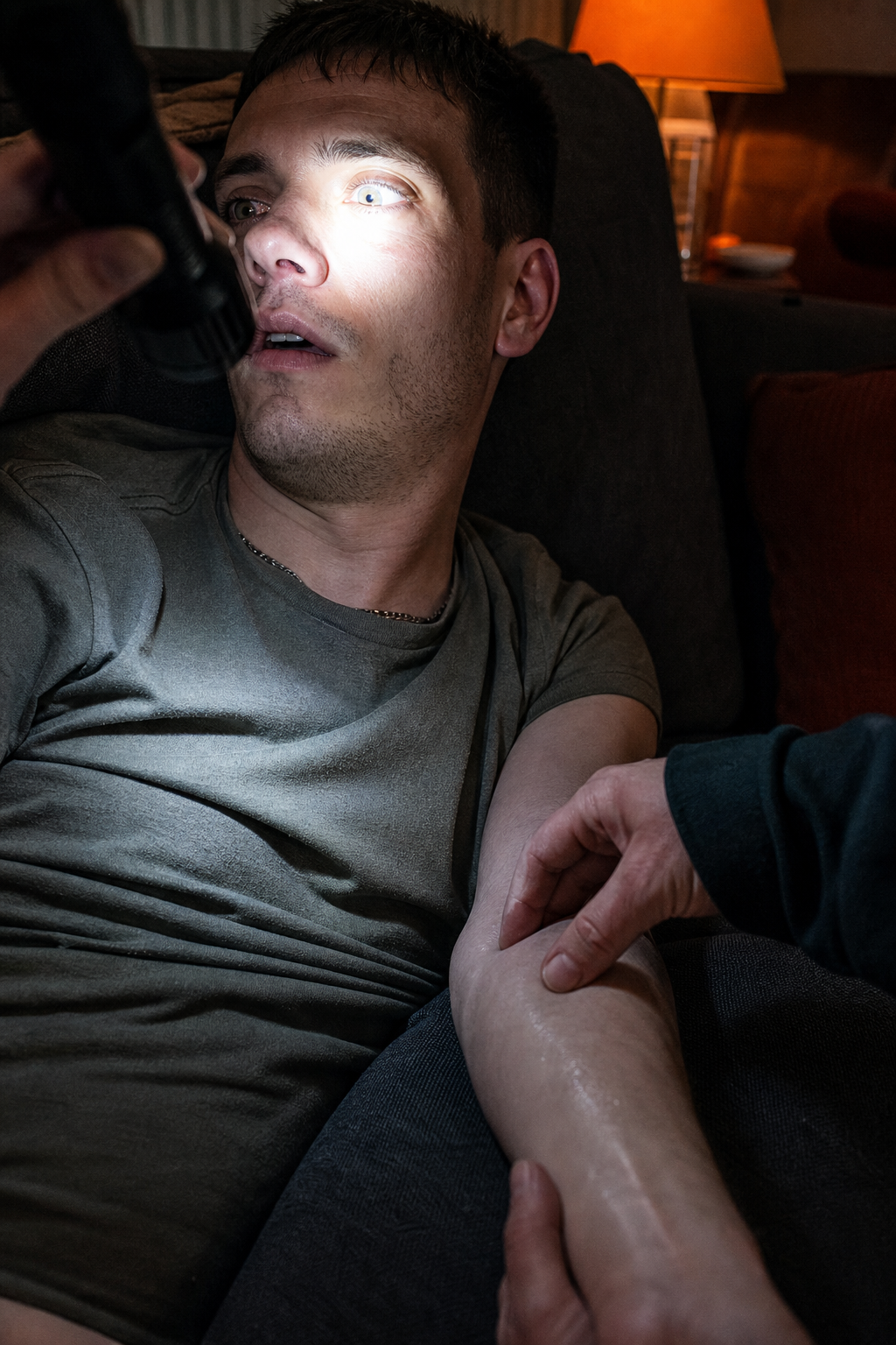 A photorealistic scene of a living room with dim lighting, a flashlight beam highlighting a person's eyes showing pinpoint pupils, and a hand feeling cool, clammy skin on the forearm. Alt: opioid overdose physical signs close‑up