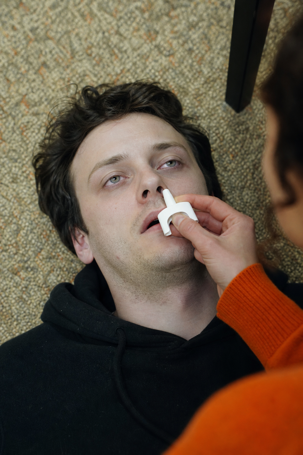 A photorealistic scene of a person's face with pale skin and tiny pupils, with another person holding a naloxone nasal spray nearby, alert expression, realistic lighting, indoor setting. Alt: Opioid overdose signs: pinpoint pupils, pale skin, unresponsiveness.