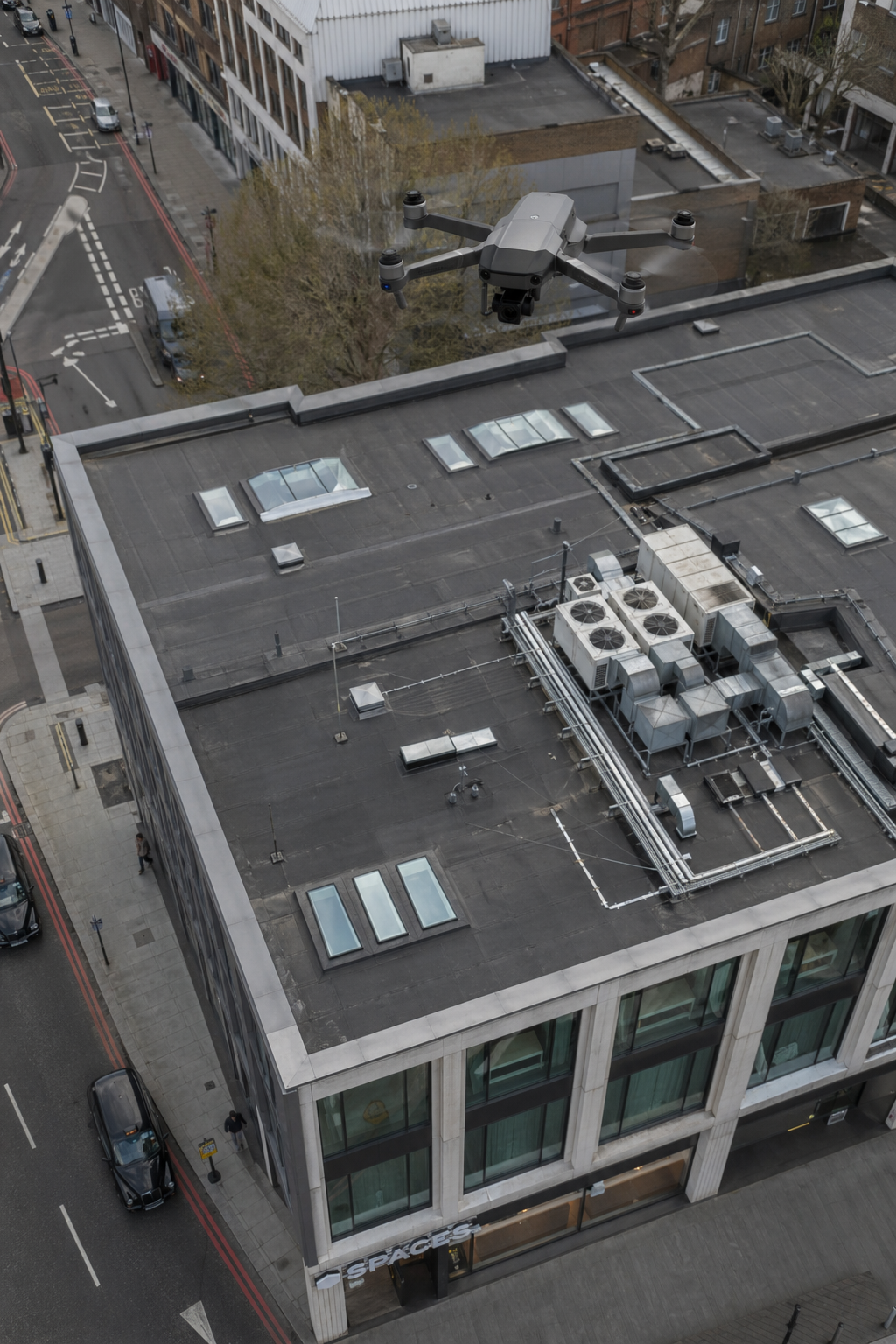 A photorealistic overhead view of a modern commercial building with a flat roof, showing a small drone hovering above, capturing high-resolution imagery. The roof has HVAC units and skylights. Alt: Drone roof inspection of commercial building.