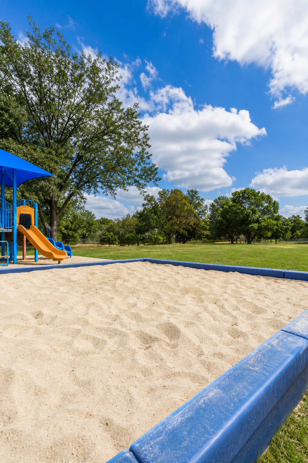 A photorealistic playground with a sandbox filled with clean, light-colored sand, surrounded by grass and trees. Texas sky, no people, no logos. Alt: Sand for playground in a San Antonio playground.