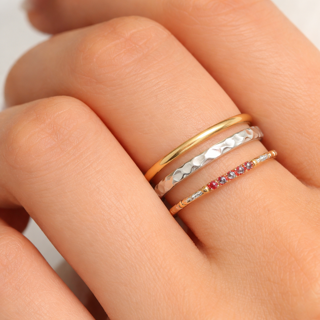 A photorealistic close‑up of a woman's hand wearing three affordable stackable rings – a thin gold‑plated band, a silver‑tone hammered ring, and a tiny ruby‑accented ring. Alt: budget-friendly stackable rings for women