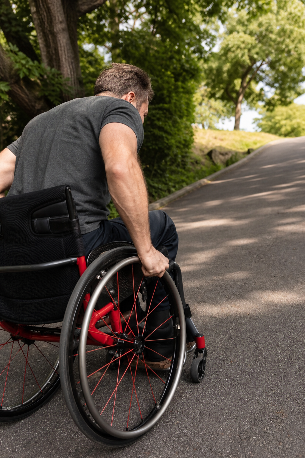 A person in a wheelchair pushing uphill on a paved path, hands gripping the hand rims with visible strain. Alt: Person propelling wheelchair uphill to demonstrate hand rim grip challenges.