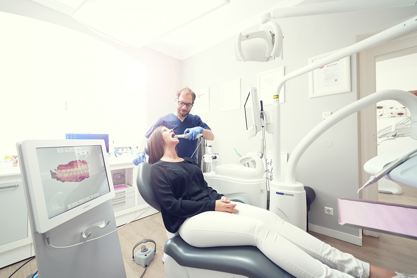 Behandlungsraum mit zurückgelehntem Dentalstuhl, zwei Personen und einem Monitor mit Gebissdarstellung.