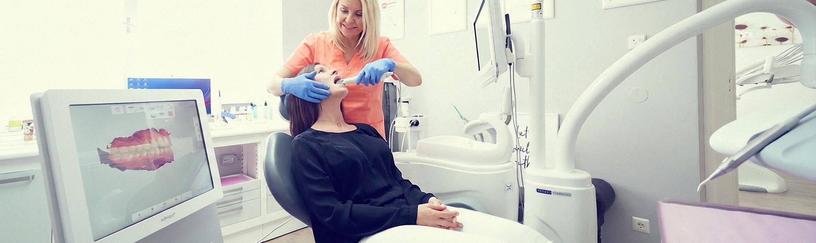 Person im Dentalstuhl und behandschuhte Hände am Mund; daneben ein Monitor mit Gebissdarstellung.
